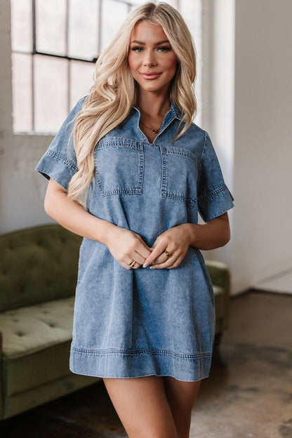 a woman wearing a denim mini dress. The dress has short sleeves, a collared neckline, and pockets.