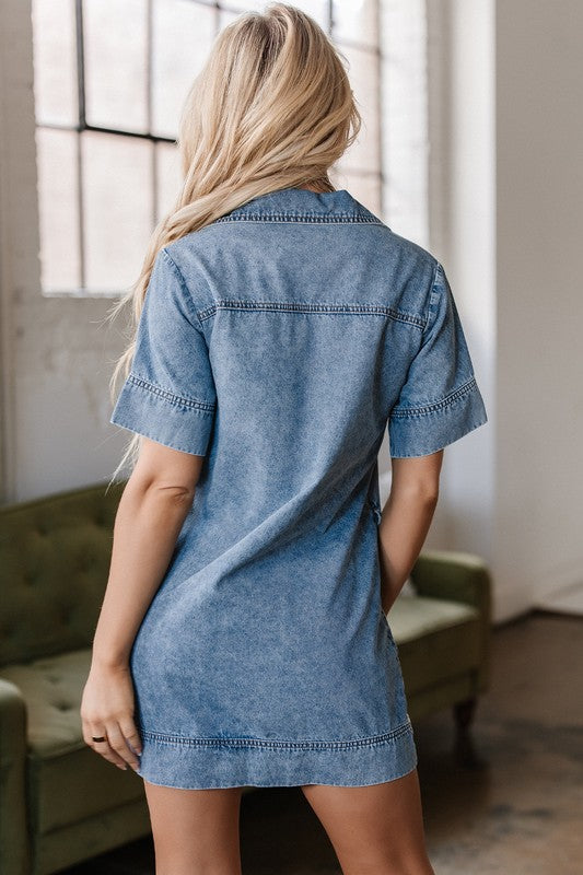 a woman wearing a denim mini dress. The dress has short sleeves, a collared neckline, and pockets.