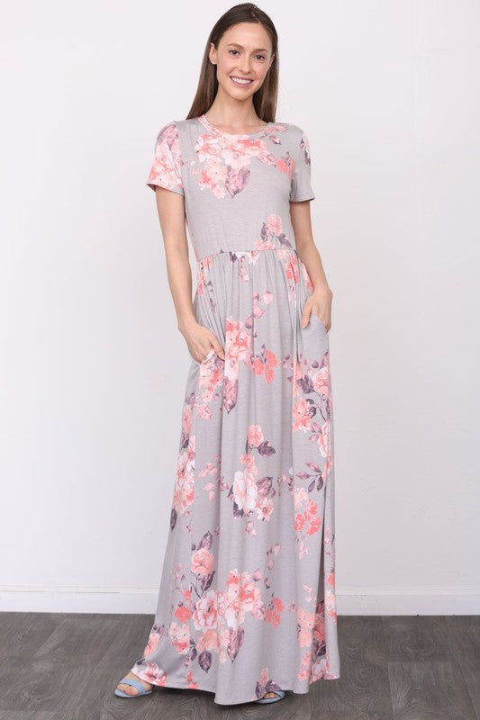 A woman in a long gray dress with pink floral patterns stands against a white wall with her hands in the dress pockets.
