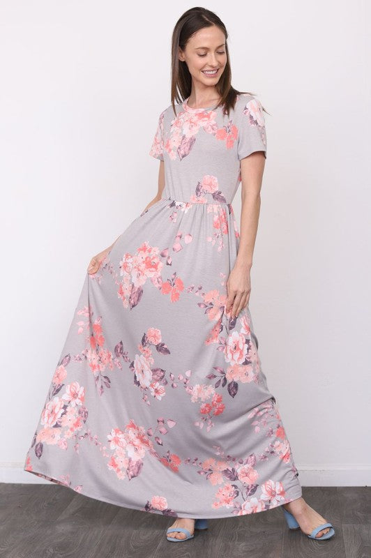 A woman in a long gray dress with pink floral patterns stands against a white wall with her hands in the dress pockets.