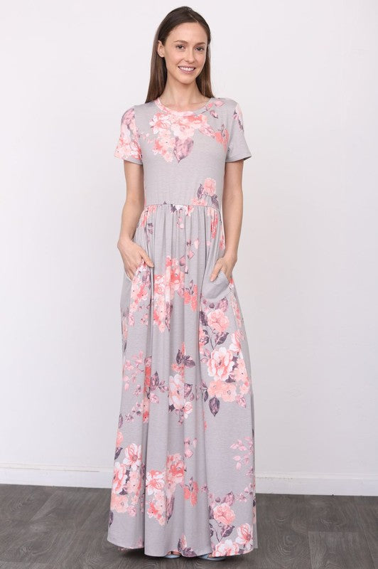 A woman in a long gray dress with pink floral patterns stands against a white wall with her hands in the dress pockets.