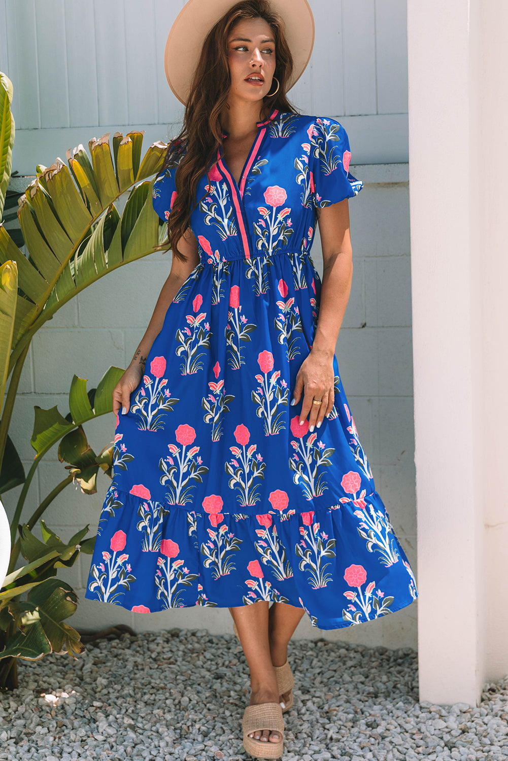 A woman posing in a vibrant blue dress with pink flowers and green leaves printed on it. She's wearing a hat, hoop earrings, and tan heels, standing on gravel next to a plant and a white wall.