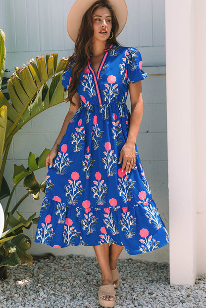 A woman posing in a vibrant blue dress with pink flowers and green leaves printed on it. She's wearing a hat, hoop earrings, and tan heels, standing on gravel next to a plant and a white wall.