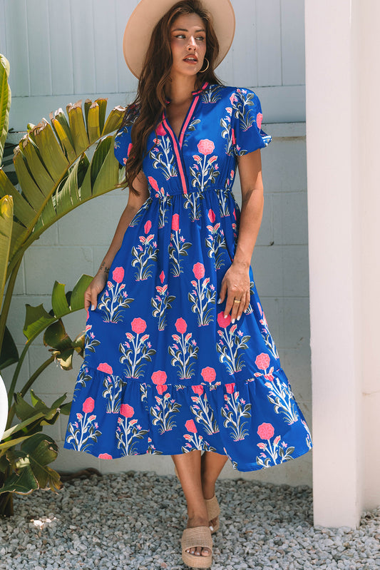 A woman posing in a vibrant blue dress with pink flowers and green leaves printed on it. She's wearing a hat, hoop earrings, and tan heels, standing on gravel next to a plant and a white wall.