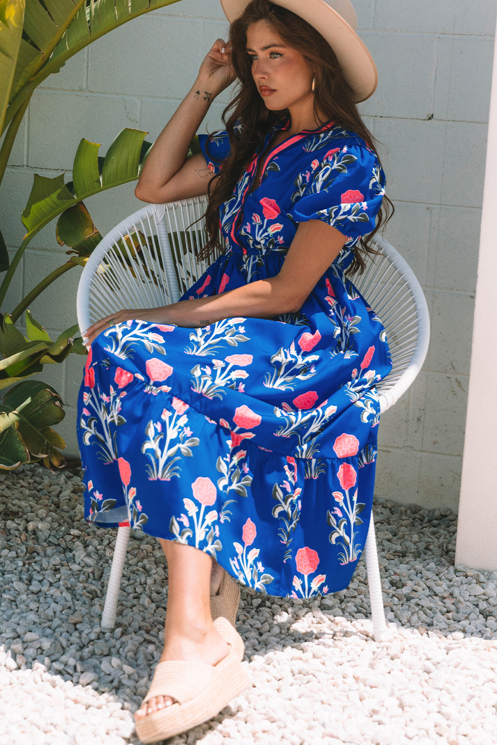 A woman posing in a vibrant blue dress with pink flowers and green leaves printed on it. She's wearing a hat, hoop earrings, and tan heels, standing on gravel next to a plant and a white wall.