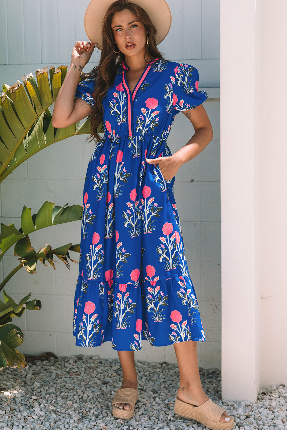 A woman posing in a vibrant blue dress with pink flowers and green leaves printed on it. She's wearing a hat, hoop earrings, and tan heels, standing on gravel next to a plant and a white wall.