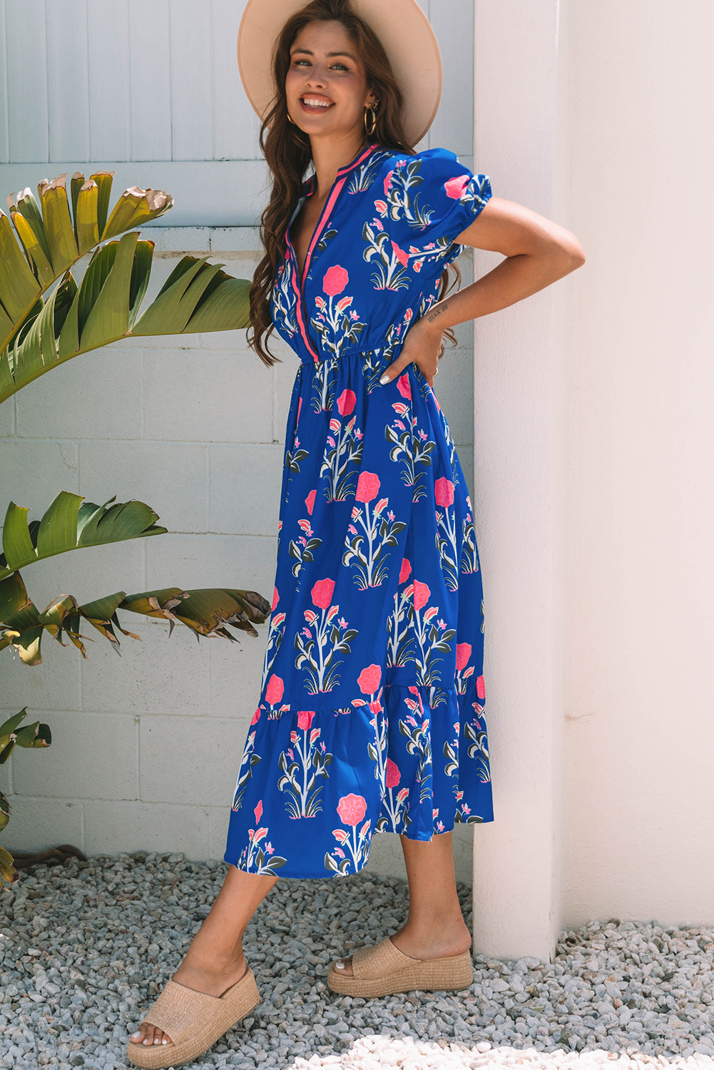 A woman posing in a vibrant blue dress with pink flowers and green leaves printed on it. She's wearing a hat, hoop earrings, and tan heels, standing on gravel next to a plant and a white wall.