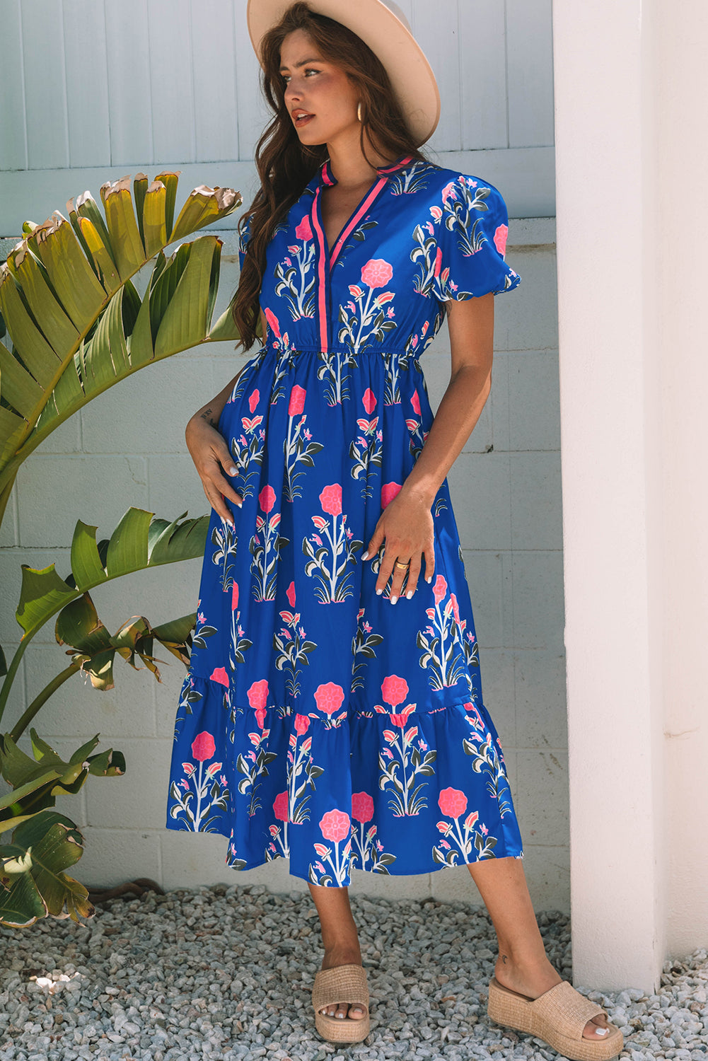 A woman posing in a vibrant blue dress with pink flowers and green leaves printed on it. She's wearing a hat, hoop earrings, and tan heels, standing on gravel next to a plant and a white wall.