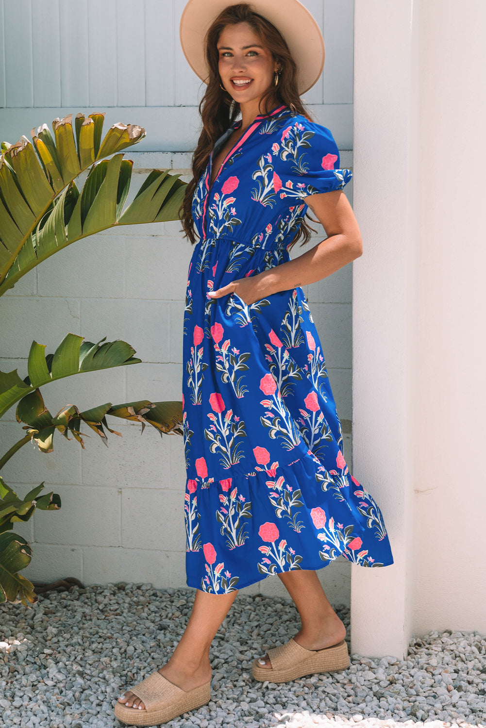 A woman posing in a vibrant blue dress with pink flowers and green leaves printed on it. She's wearing a hat, hoop earrings, and tan heels, standing on gravel next to a plant and a white wall.