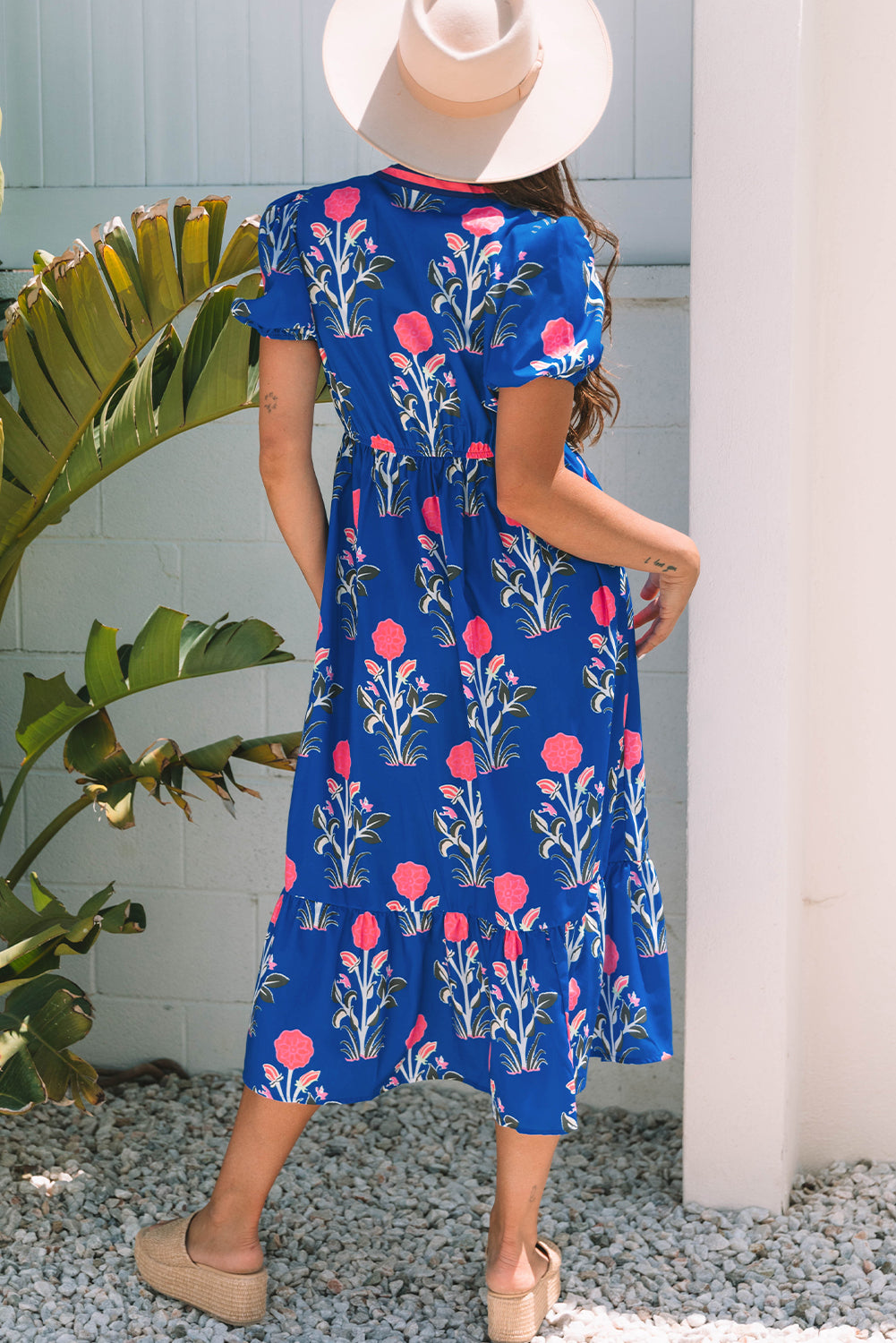 A woman posing in a vibrant blue dress with pink flowers and green leaves printed on it. She's wearing a hat, hoop earrings, and tan heels, standing on gravel next to a plant and a white wall.