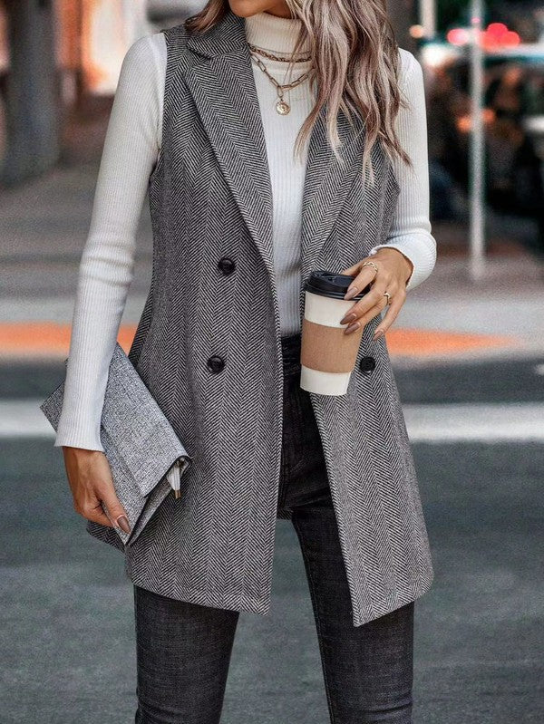 A woman wearing a long gray vest over a white turtleneck and dark pants. She's holding a coffee cup in one hand and a clutch in the other. She's standing on a street with a blurred background.
