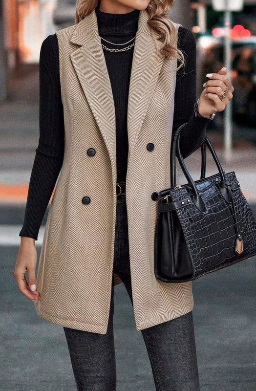 A woman wearing a long beige vest over a black turtleneck and gray pants, holding a black handbag.
