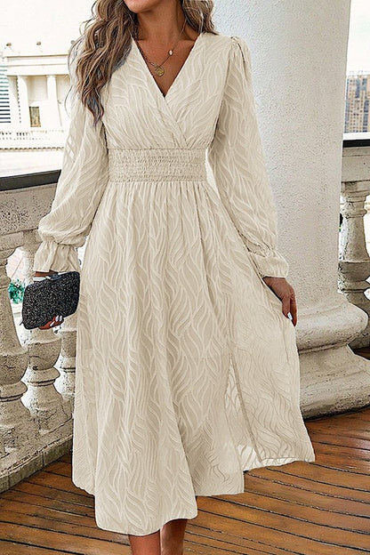 Woman wearing a cream long sleeve midi dress with a smocked waist and flowing skirt, styled with a clutch on a balcony setting.