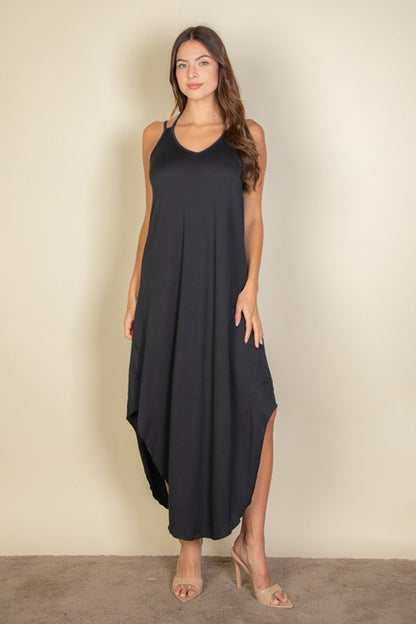 A woman with long brown hair is posing in a black, sleeveless midi dress with a V-neck and a slit on one side. She's wearing tan heels and standing on a beige carpet against a beige wall.