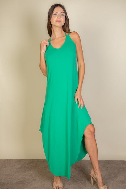 A woman with long brown hair is posing in a green, sleeveless midi dress with a V-neck and a slit on one side. She's wearing tan heels and standing on a beige carpet against a beige wall 