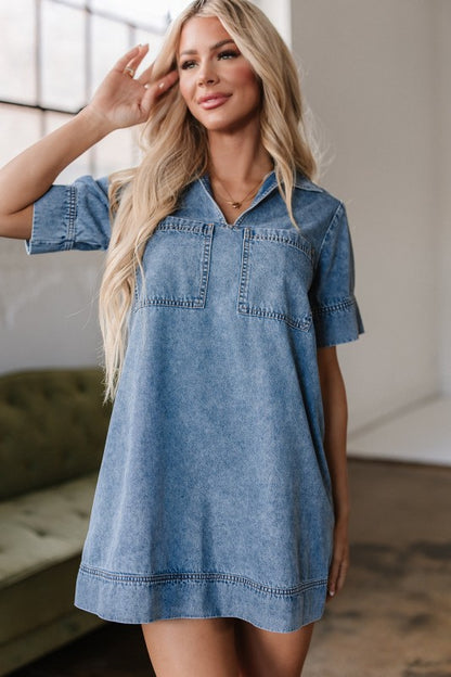 A young woman poses in a short-sleeved, collared denim mini dress in faded blue wash, featuring a V-neckline, front patch pockets