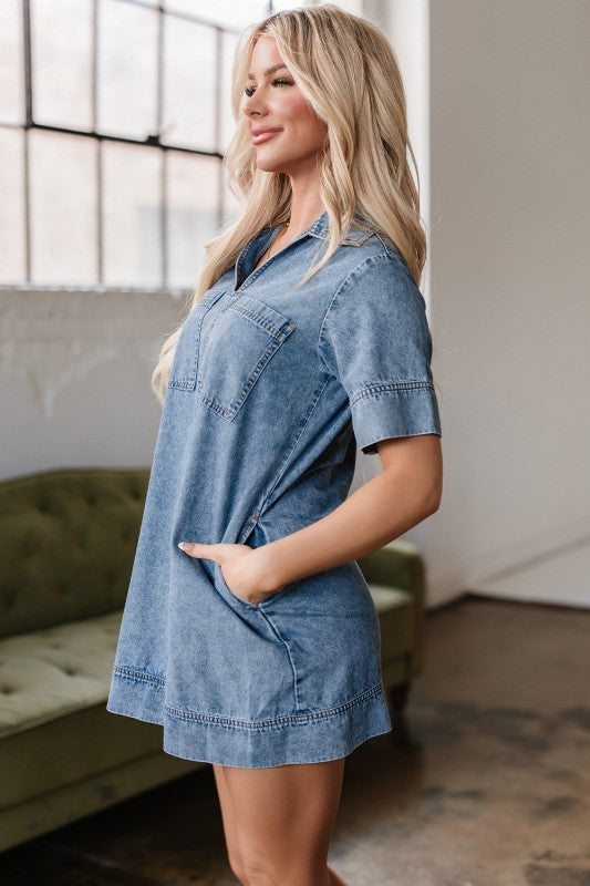A young woman poses in a short-sleeved, collared denim mini dress in faded blue wash, featuring a V-neckline, front patch pockets