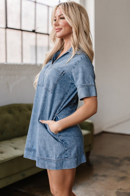 A young woman poses in a short-sleeved, collared denim mini dress in faded blue wash, featuring a V-neckline, front patch pockets