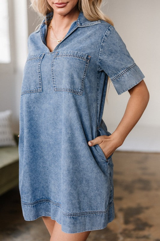A young woman poses in a short-sleeved, collared denim mini dress in faded blue wash, featuring a V-neckline, front patch pockets