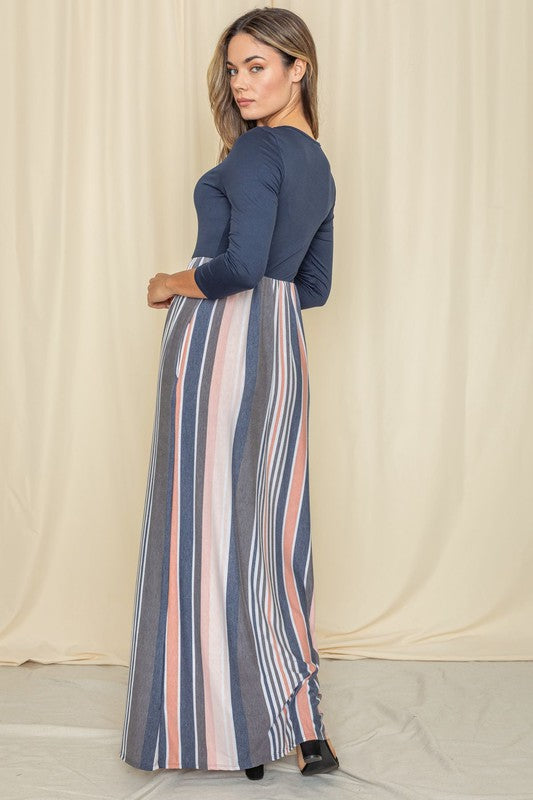 Woman in a navy blue top with a striped maxi skirt (blue, pink, white) posing against a cream curtain backdrop.