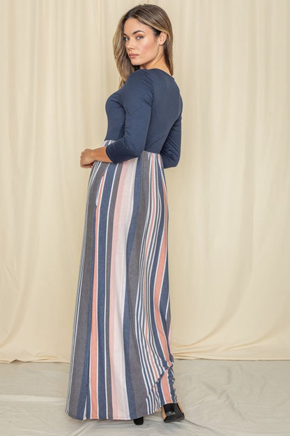 Woman in a navy blue top with a striped maxi skirt (blue, pink, white) posing against a cream curtain backdrop.
