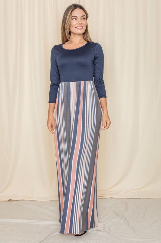 Woman in a navy blue top with a striped maxi skirt (blue, pink, white) posing against a cream curtain backdrop.