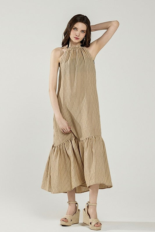 A young woman posing in a tan striped sleeveless dress with a ruffled hem and tan sandals against a plain background.
