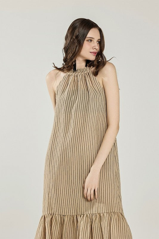 A young woman posing in a tan striped sleeveless dress with a ruffled hem and tan sandals against a plain background.