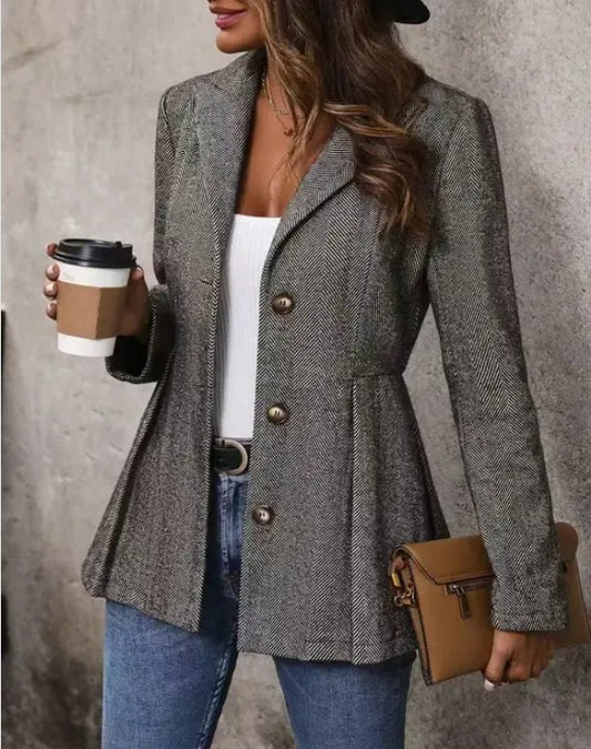  A woman wearing a gray tweed blazer with brass buttons, a white top, blue jeans, and a black hat. She's holding a coffee cup in one hand and a brown purse in the other, posing against a gray wall.