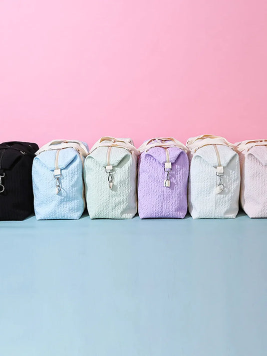 A row of seven quilted backpacks in various colors—black, light blue, mint green, lavender, white, and light pink—neatly arranged against a pastel pink and blue background. Uniform design with flap closures and top handles—stylish product lineup.