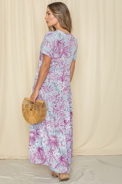 A woman in a floral maxi dress with short sleeves, standing against a cream curtain backdrop.