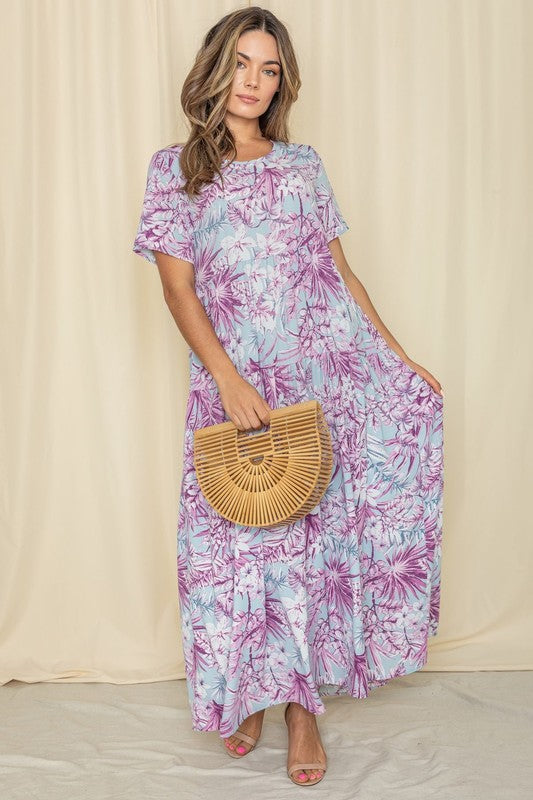 A woman posing in a long, flowy lilac and purple floral dress with short sleeves, holding a half-moon wandy purse in front of a cream curtain backdrop. 