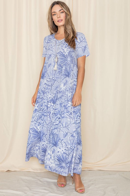 A woman in a blue floral maxi dress with short sleeves, standing against a cream curtain backdrop.