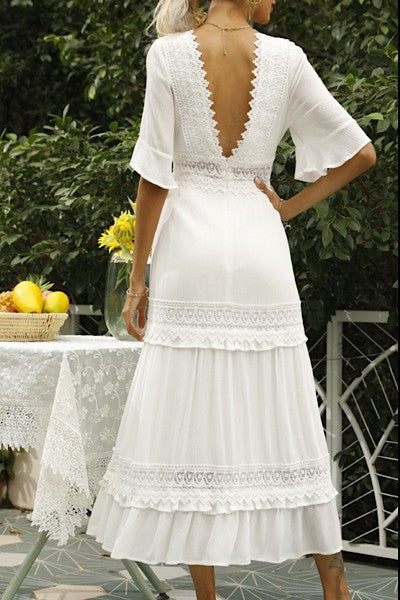 A woman in a white dress . She's standing sideways, looking towards the camera with a subtle smile. The dress is long, has lace details, and she's rocking white heels. Background's plain light grey.