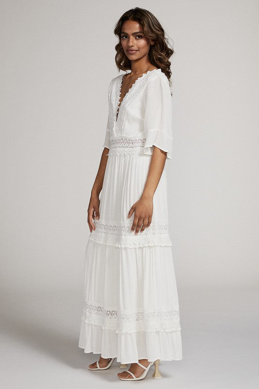A woman in a white dress . She's standing sideways, looking towards the camera with a subtle smile. The dress is long, has lace details, and she's rocking white heels. Background's plain light grey.