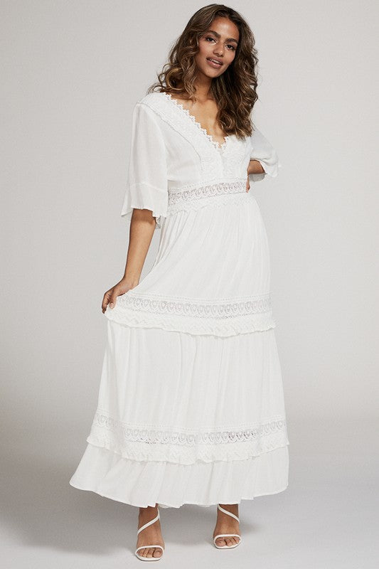 A woman in a white dress . She's standing sideways, looking towards the camera with a subtle smile. The dress is long, has lace details, and she's rocking white heels. Background's plain light grey.