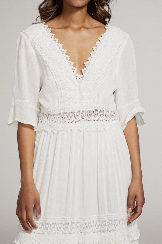 A woman in a white dress . She's standing sideways, looking towards the camera with a subtle smile. The dress is long, has lace details, and she's rocking white heels. Background's plain light grey.
