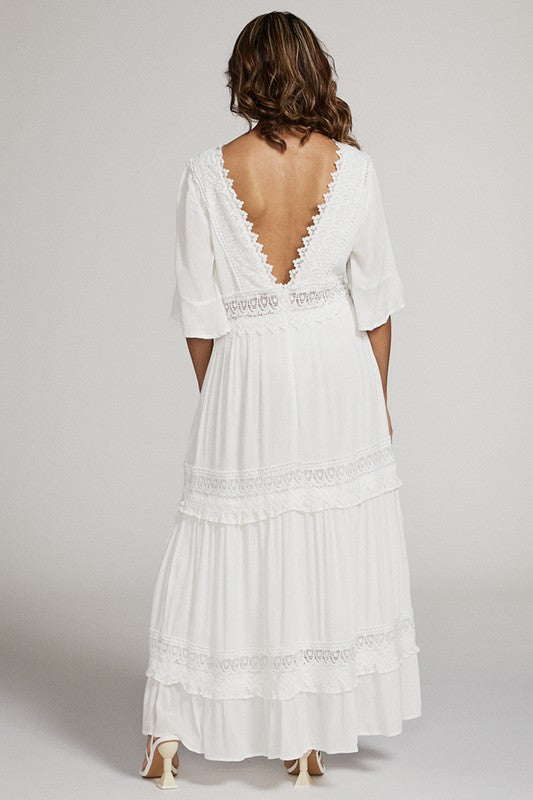 A woman in a white dress . She's standing sideways, looking towards the camera with a subtle smile. The dress is long, has lace details, and she's rocking white heels. Background's plain light grey.