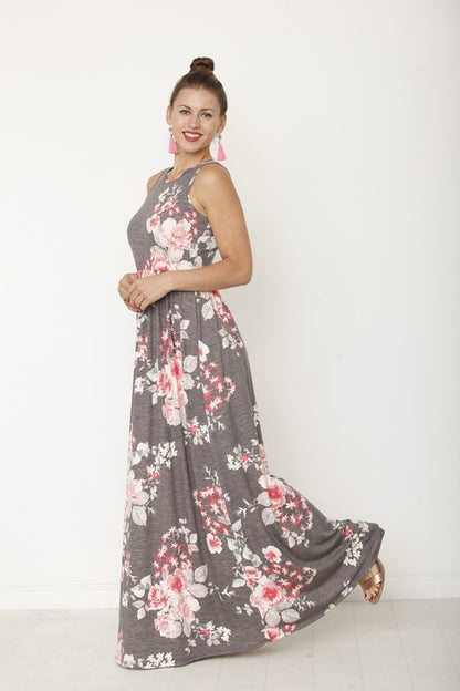A woman in a long, gray sleeveless dress with a floral pattern of pink and white flowers. She's standing against a white wall, with her hair in a bun and wearing pink earrings and tan sandals. 