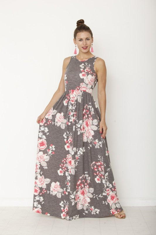 A woman in a long, gray sleeveless dress with a floral pattern of pink and white flowers. She's standing against a white wall, with her hair in a bun and wearing pink earrings and tan sandals. 