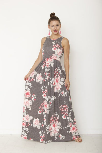 A woman in a long, gray sleeveless dress with a floral pattern of pink and white flowers. She's standing against a white wall, with her hair in a bun and wearing pink earrings and tan sandals. 