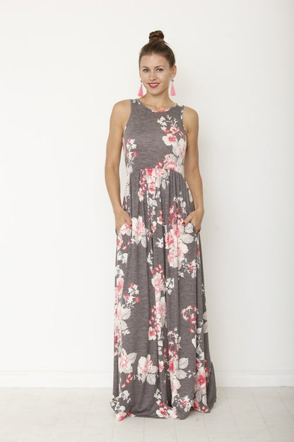 A woman in a long, gray sleeveless dress with a floral pattern of pink and white flowers. She's standing against a white wall, with her hair in a bun and wearing pink earrings and tan sandals. 