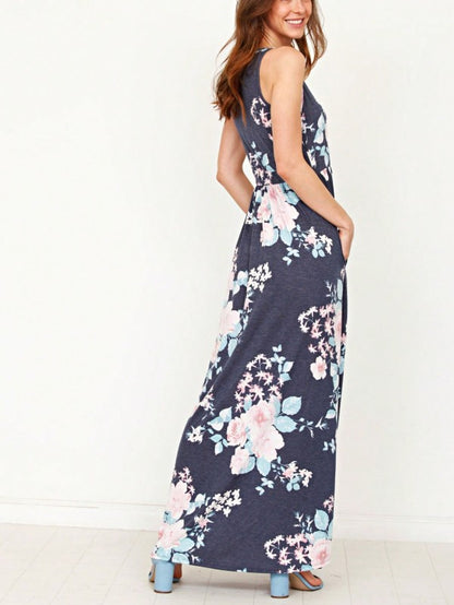 A woman in a long, gray sleeveless dress with a floral pattern of pink and white flowers. She's standing against a white wall, with her hair in a bun and wearing pink earrings and tan sandals. 