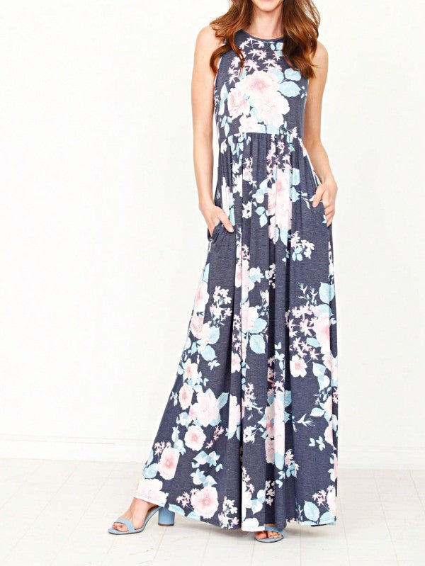 A woman in a long, gray sleeveless dress with a floral pattern of pink and white flowers. She's standing against a white wall, with her hair in a bun and wearing pink earrings and tan sandals. 