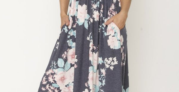 A woman in a long, gray sleeveless dress with a floral pattern of pink and white flowers. She's standing against a white wall, with her hair in a bun and wearing pink earrings and tan sandals. 