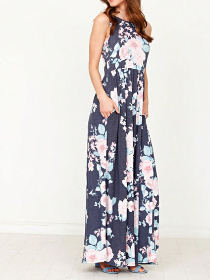 A woman in a long, sleeveless dress with a floral pattern of pink and white flowers. She's standing against a white wall, with her hair in a bun and wearing pink earrings and tan sandals. 
