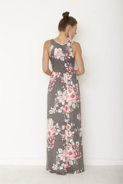 A woman in a long, gray sleeveless dress with a floral pattern of pink and white flowers. She's standing against a white wall, with her hair in a bun and wearing pink earrings and tan sandals. 