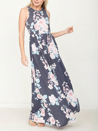 A woman in a long, gray sleeveless dress with a floral pattern of pink and white flowers. She's standing against a white wall, with her hair in a bun and wearing pink earrings and tan sandals. 