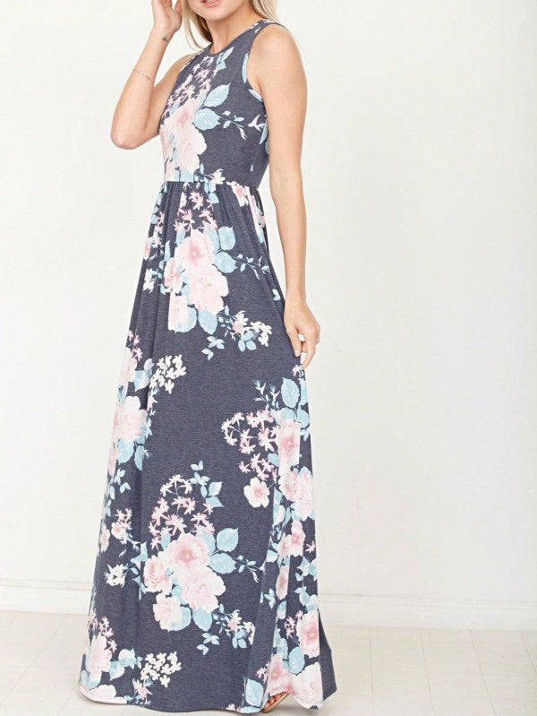 A woman in a long, gray sleeveless dress with a floral pattern of pink and white flowers. She's standing against a white wall, with her hair in a bun and wearing pink earrings and tan sandals. 