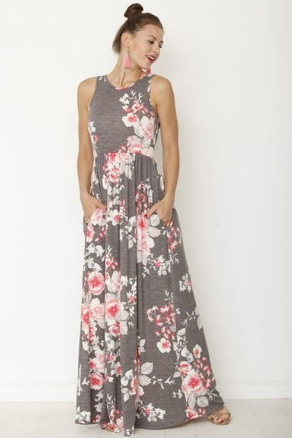 A woman in a long, gray sleeveless dress with a floral pattern of pink and white flowers. She's standing against a white wall, with her hair in a bun and wearing pink earrings and tan sandals. 